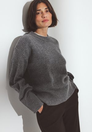 Gray knit sweater with a crew neck, ribbed cuffs and hem, made from soft material; worn over dark trousers against a plain wall.