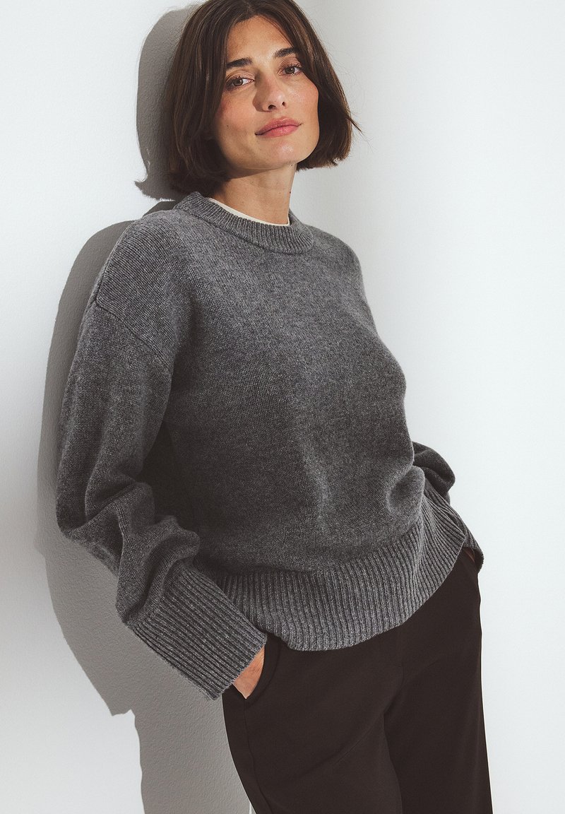 Gray knit sweater with a crew neck, ribbed cuffs and hem, made from soft material; worn over dark trousers against a plain wall.