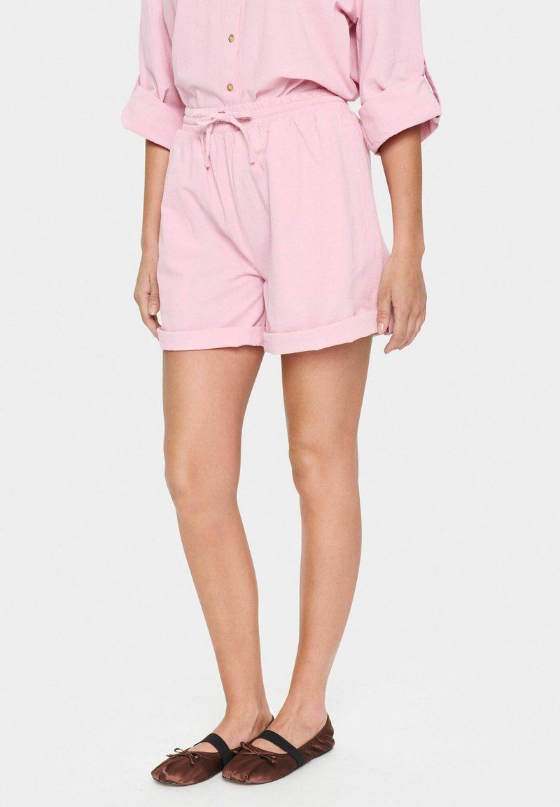 Pantaloni corti di cotone rosa chiaro con vita elastica, lacci e orli arrotolati. Abbinati a scarpe basse marroni con un accento a fiocco.