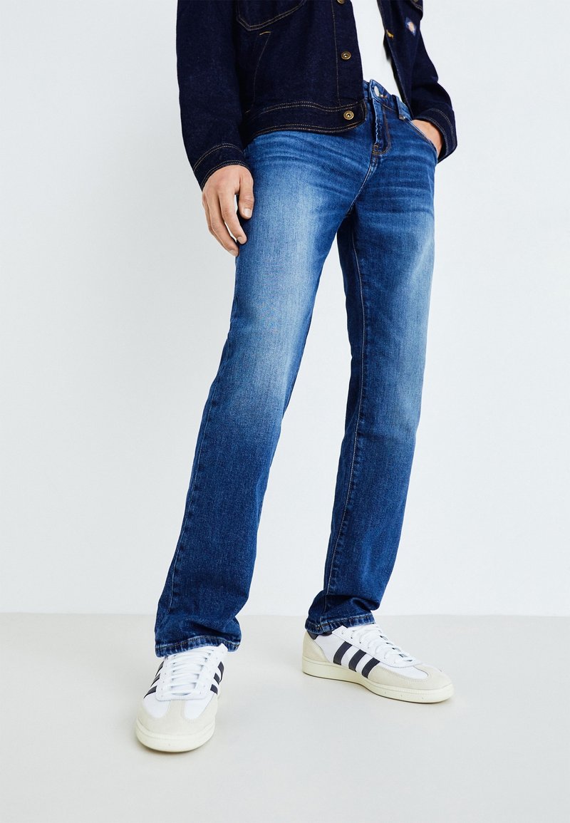 Ljusblå denimjeans med avslappnad passform och blekt utseende, kombinerat med vita sneakers med svarta ränder.