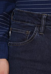 Tmavé džínové kalhoty s klasickým střihem, kontrastním šitím a malým logem na přední kapse. Hladká textura a tradiční design.