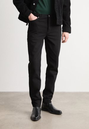 Homme portant un pantalon noir, des chaussures en cuir noires, une veste sombre et une chemise verte, debout sur un sol gris avec une main dans la poche.