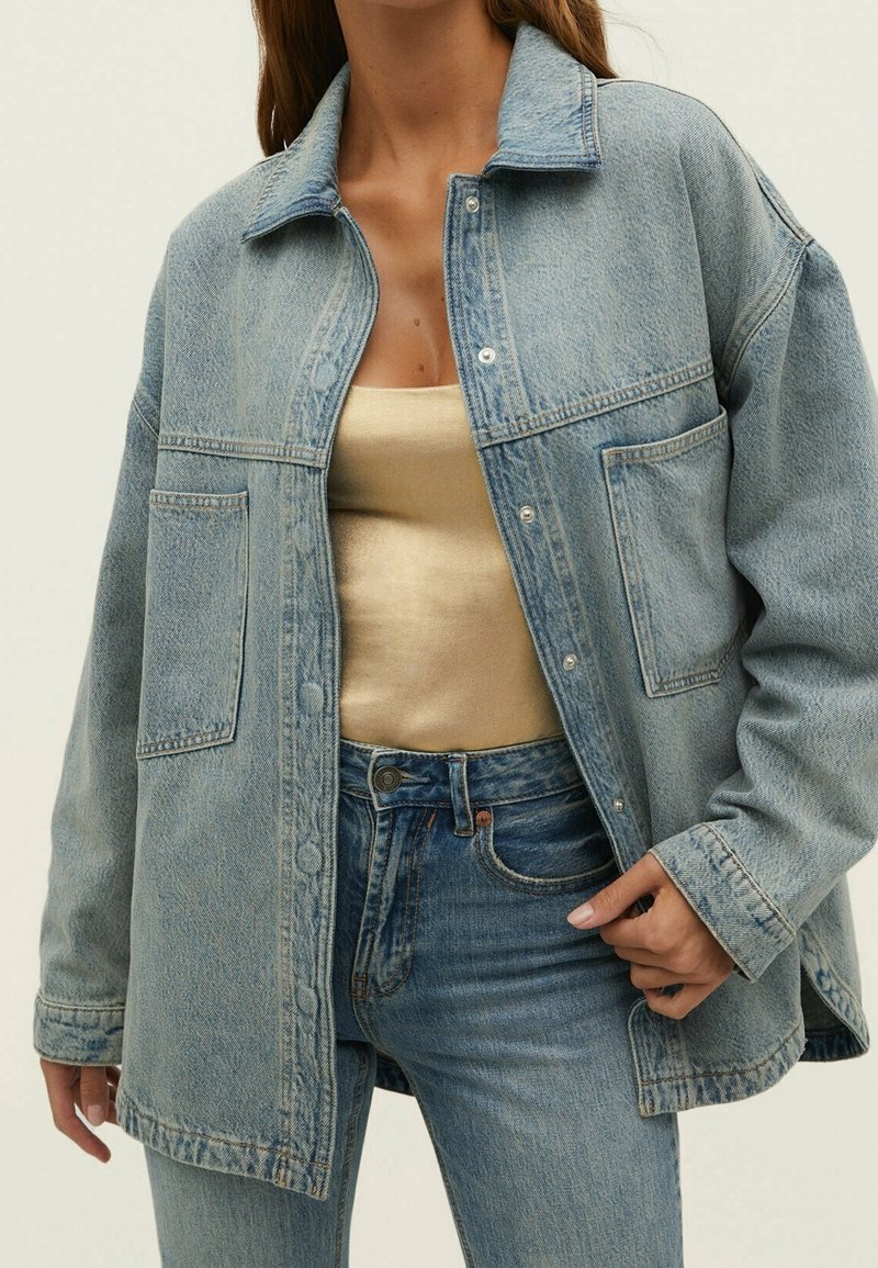 Vrouw in een lichtblauwe denimjas over een gouden satijnen top en blauwe denim jeans, hand die de jas bij de taille vasthoudt.