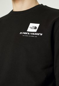 Svart bomullssweatshirt med en vit logotyp och geografiska koordinater på bröstet. Ribbad halsringning och en slät textur genomgående.