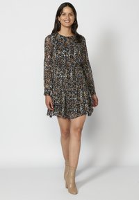 Schwarzes Blumenmaxikleid mit langen Ärmeln und einem Rüschensaum, das mehrfarbige abstrakte Muster zeigt. Kombiniert mit beigen Absatzstiefeln.
