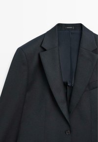 Blazer tailored blu navy con revers a ribalta, chiusura a un bottone e una texture sottile. Presenta una fodera interna liscia e una tasca sul petto.