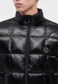 Homme portant une doudoune matelassée noire brillante avec un col montant et un logo rond Armani Exchange sur la poitrine.