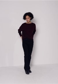 Pull bordeaux à manches longues, coupe décontractée, associé à un jean noir slim. Le mannequin se tient devant un fond neutre.