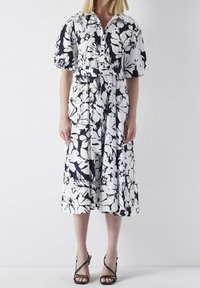 Blommönstrad klänning i svart och vitt, med krage, puffärmar, ett insvängt midja, och en flödande kjol som slutar vid vaderna.