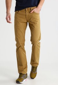 Beige bomulls chinos med rak passform, utrustade med fem fickor och diskreta sömmar. Bärs med sportskor i grått och grönt.