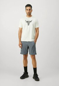 Lysefarvet t-shirt med et sort bulleskalle-grafik, parret med grå shorts og sorte træningssko, stående på en ensfarvet baggrund.