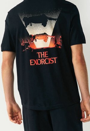 T-shirt noir avec un graphisme d'une fille lévitante et deux figures sous une lumière vive, étiqueté "The Exorcist" en texte rouge.