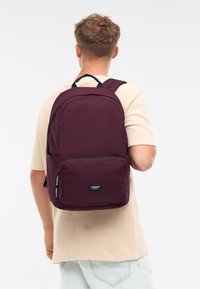 Rucsac burgundy cu curele căptușite, buzunar frontal cu fermoar și patch cu logo. Fabricat dintr-un material rezistent, cu o textură fină și un design structural.