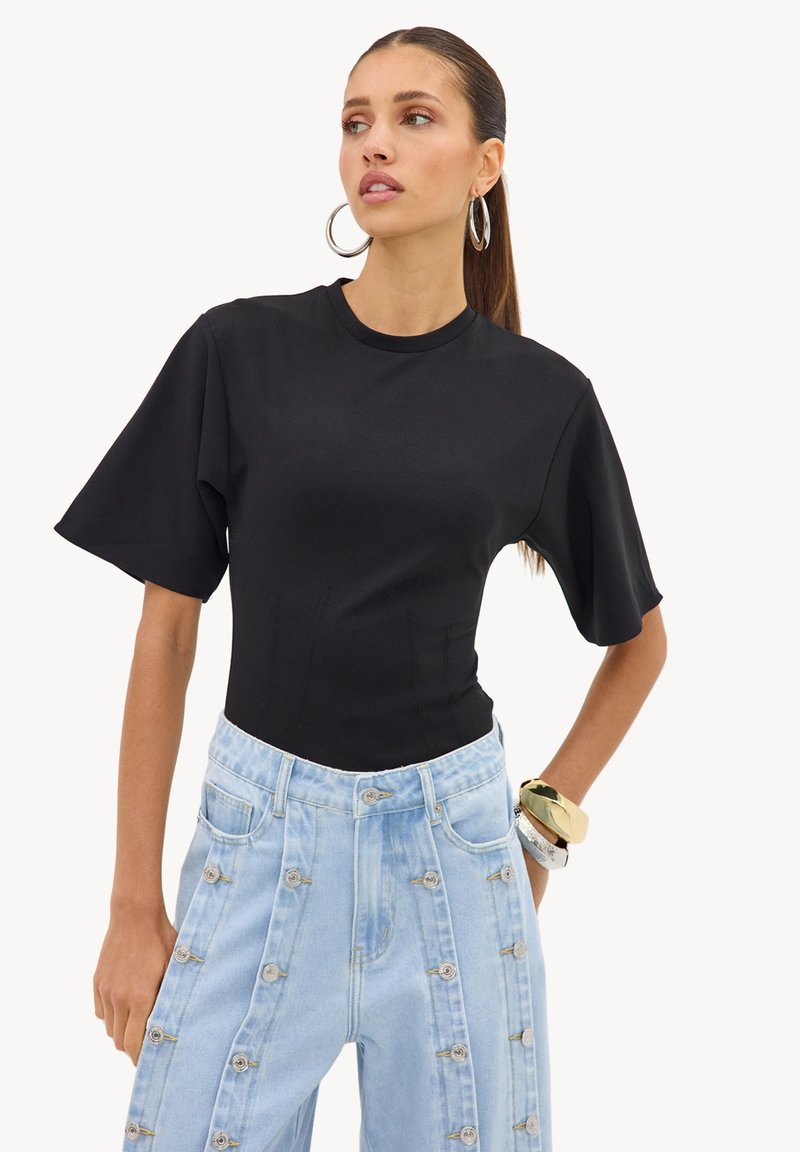 Femme aux cheveux foncés en queue de cheval portant un haut noir à manches courtes, un jean bleu clair taille haute avec des boutons décoratifs, de grandes boucles d'oreilles et des bracelets.
