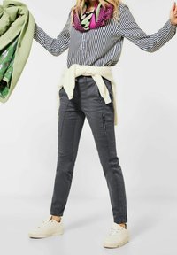 Grå slim-fit byxor med dragkedjefickor och sidodetaljer, kombinerat med en randig skjorta och en färgglad scarf, bärs med vita sneakers.