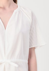 Weiße Bluse mit V-Ausschnitt, gerafftem Taillenzug und semi-transparenten, strukturierten Kurzärmeln an einer Person vor einem schlichten Hintergrund.