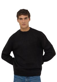 Czarny sweter dzianinowy z okrągłym dekoltem, ściągaczami przy mankietach i u dołu, oraz luźnym krojem. Noszony z niebieskimi dżinsami.