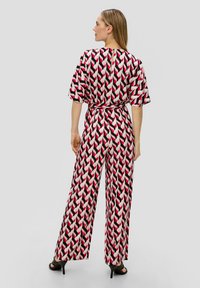 Geprinte jumpsuit met een gedurfde geometrische patroon in roze, rood en zwart. Wijde benen, losse pasvorm en een ceintuur met een keyhole rug.