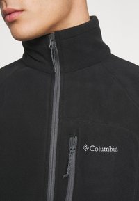 Svart fleecejakke med høy krage, frontlomme med glidelås og brystlomme. Har en grå glidelås og Columbia-logo på venstre side.