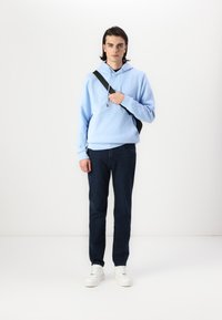 Hanorac albastru deschis cu buzunar tip cangur, curea neagră pe umăr, blugi negri din denim și adidași albi cu tălpi texturate. Design minimalist.