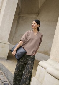 Blusa marrone a trama con scollo a V e bordo arricciato, abbinata a pantaloni larghi neri con motivi a foglia d'oro. In mano tiene una pochette nera.
