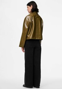 Sieviete, kura stāv ar muguru, tērpusies olīvzaļā ādas jakā un platiem melniem džinsiem uz balta fona.