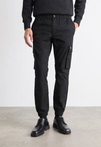 Pantaloni cargo negri dintr-un material rezistent, cu talie elastică, manșete înguste, buzunare laterale și o etichetă cu logo negru.