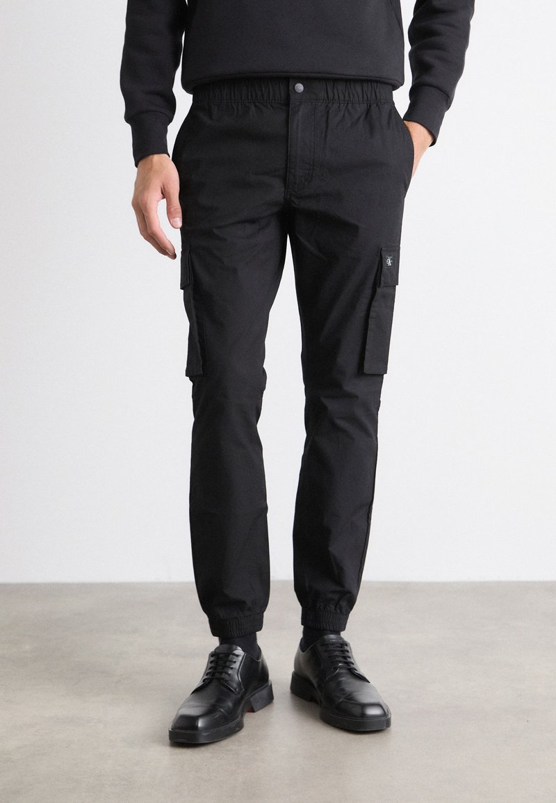 Pantaloni cargo negri dintr-un material rezistent, cu talie elastică, manșete înguste, buzunare laterale și o etichetă cu logo negru.