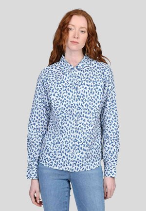 Chemise blanche à boutons avec un motif floral bleu, manches longues et col pointu. Le modèle l'associe avec un jean bleu clair.