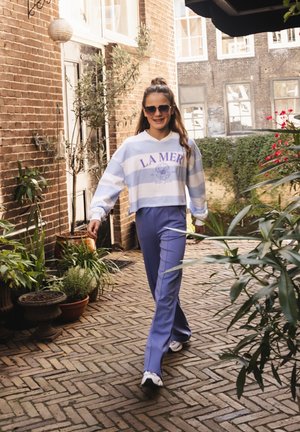 Young woman in sunglasses walking in a courtyard wearing a striped "LA MER" sweatshirt and blue pants surrounded by potted plants.