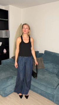 Γυναίκα με μαύρο αμάνικο τοπ, μπλε τζιν παντελόνι με φαρδιά μπατζάκια και μαύρες γόβες, στέκεται στο σαλόνι κρατώντας μικρό μαύρο τσαντάκι.