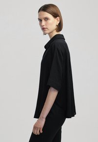 Blusa nera a maniche corte con colletto, caratterizzata da una vestibilità rilassata e una texture liscia, abbinata a pantaloni neri aderenti.