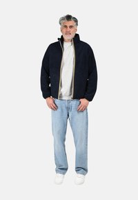 Giacca con zip color navy con finitura testurizzata, cerniera oro e bianca, abbinata a una felpa grigia, jeans azzurri e sneakers bianche.