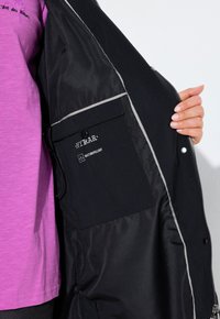 Personne tenant une veste noire ouverte montrant une poche intérieure étiquetée "HYRAR" et "imperméable" avec un bord réfléchissant à l'intérieur de la veste.