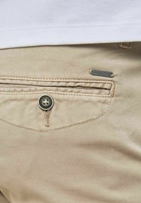 Beige bavlněné kalhoty s přední kapsou na knoflík, zvýrazněné kovovým štítkem "JACK & JONES" připevněným na boku.