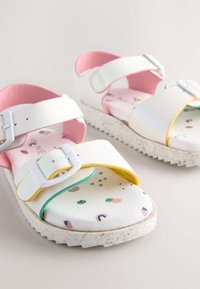 Weiße Sandalen mit rosa und gelben Akzenten, ausgestattet mit einer strukturierten Sohle und bunten Fruchtmustern auf den Einlegesohlen. Verstellbare Riemen.