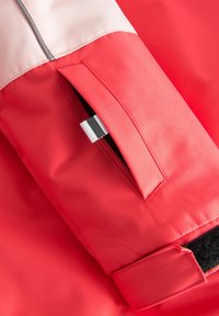 Röd tyg med en jämn textur har en dragkedjeficka och kardborrem. Den kontrasterande övre sektionen är ljusare, med grå kant.