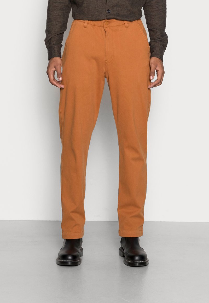 Calças em laranja enferrujado, feitas de mistura de algodão. Design de perna reta, com passantes para cinto e bolsos laterais. Botas de tornozelo pretas completam o look.