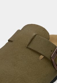 Nahaufnahme einer olivgrünen Wildledersandale mit einem breiten Riemen und einer bronzefarbenen Metallschnalle.