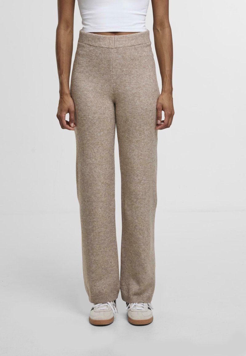 Béžové pletené kalhoty s širokými nohavicemi a vroubkovaným pasem. Měkká textura a uvolněný střih, doplněné světlými teniskami.