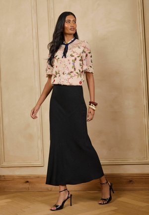 Frau trägt eine florale Spitzenbluse mit einer marineblauen Schleife, schwarze Knöchelriemchenabsätze, goldene und braune Armreifen und einen langen schwarzen Rock und steht drinnen.