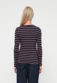 Une femme aux longs cheveux blonds, portant un haut à rayures bleu marine et marron à manches longues et un jean bleu foncé, de dos, sur fond blanc.