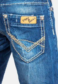 Denim jeans in een vervaagde blauwe kleur, met contrasterende gele stiksels en een leren label met "Cipo & Baxx" op de achterzak.
