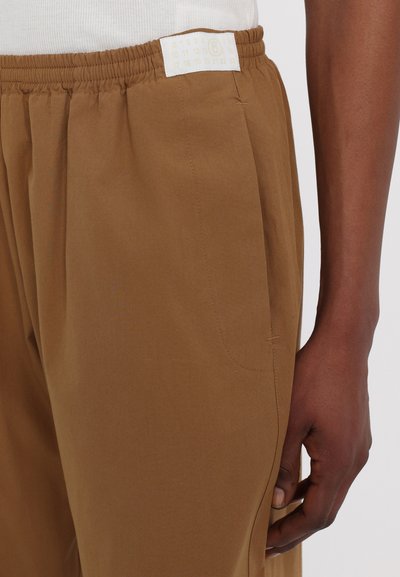 Brune bukser med elastisk talje, med en sidelomme og et etikett med numerisk design. Materialet er glat med en let glans.