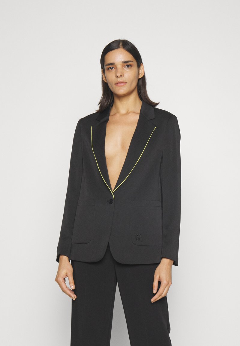 Crni sako s neonskim žutim obrubom, zatvaranje na jedan gumb, dva prednja džepa i diskretna vezenina loga na džepu. Glatka tekstura tkanine.