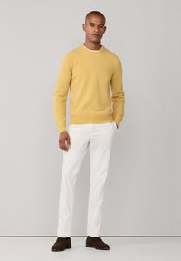 Pull en laine jaune avec col rond, trousers blancs et chaussures en suède marron. Design simple, tissu texturé, silhouette ajustée.