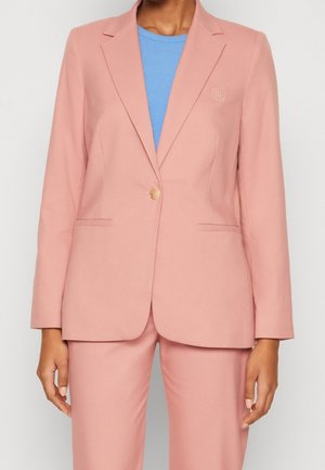 Vrouw draagt een op maat gemaakt lichtroze pak met één knoop en daaronder een lichtblauw T-shirt met een ronde hals.