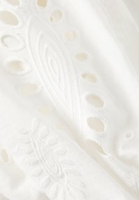 Valkoinen kangas, jossa on monimutkaista kirjaillua lehti- ja kukkakuviota. Pieniä pyöreitä aukkoja korostaa muotoilua. Pehmeä tekstuuri.