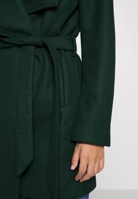 Grön kappa med en strukturerad finish, bälte i midjan och långa ärmar. Bältet är knutet fram, vilket kompletterar den övergripande designen.