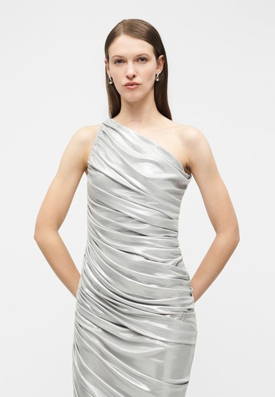Γυναίκα που φοράει γυαλιστερό ασημί φόρεμα με έναν ώμο και ρίγες, με ίσια μαλλιά και κρίκους στα αυτιά, στέκεται μπροστά σε λευκό σκηνικό.